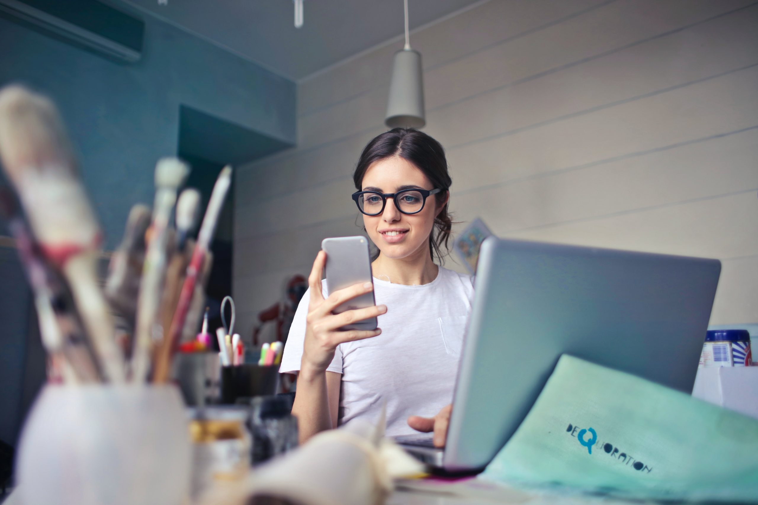 Woman wearing glasses checking on her laptop and phone messages surrounded by art supplies.