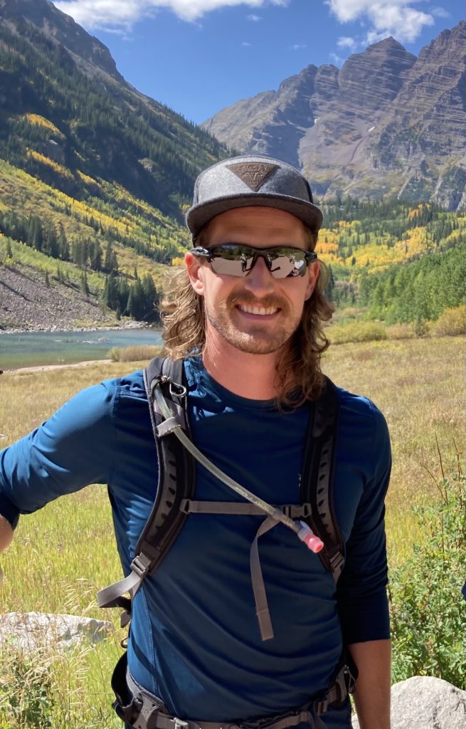 Man wearing sunglasses standing in the grass with mountains and trees in background.