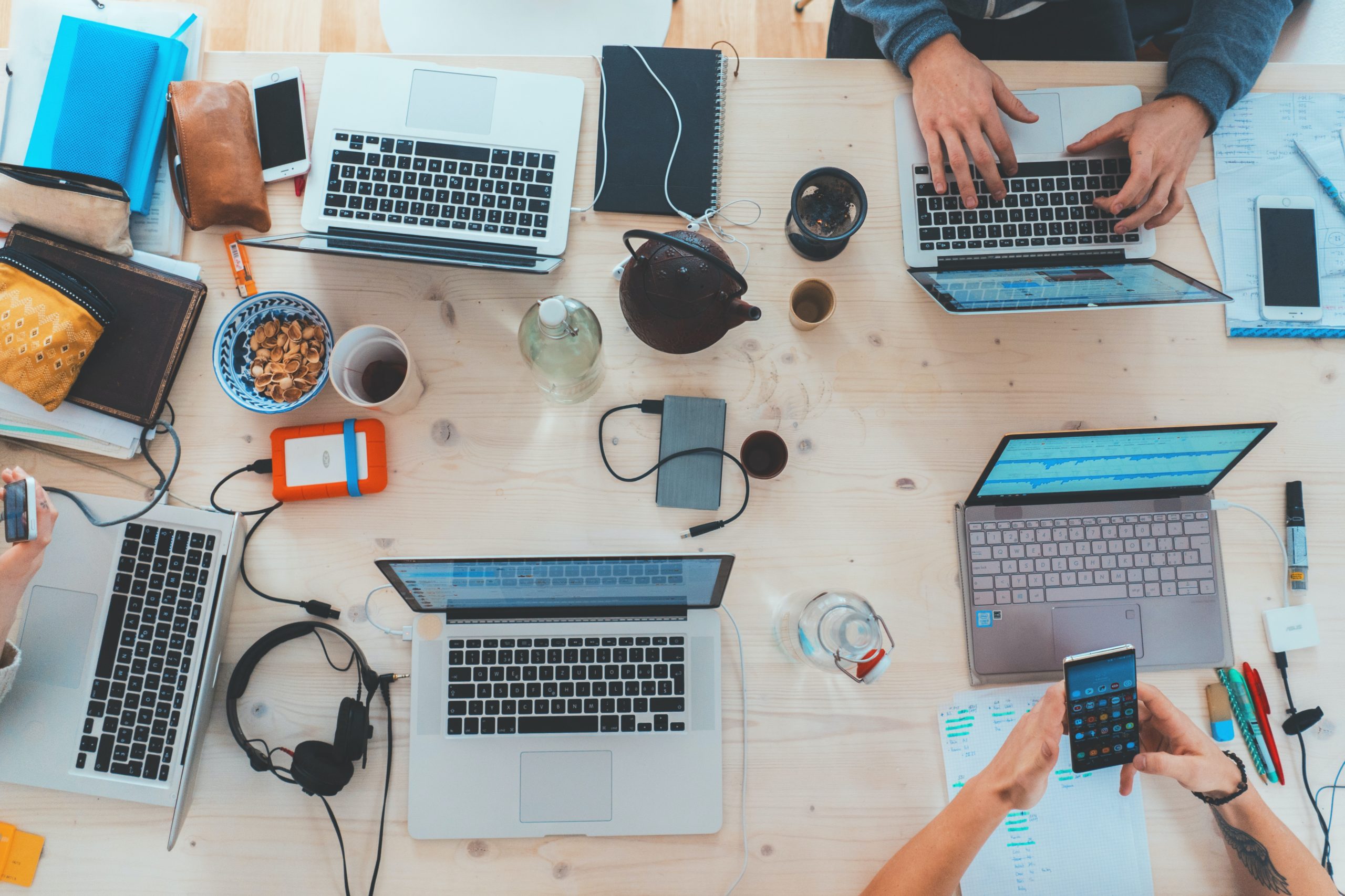 5 laptops gathered at a table with coffee mugs, bowls of snacks, headphones, and phones beside them.